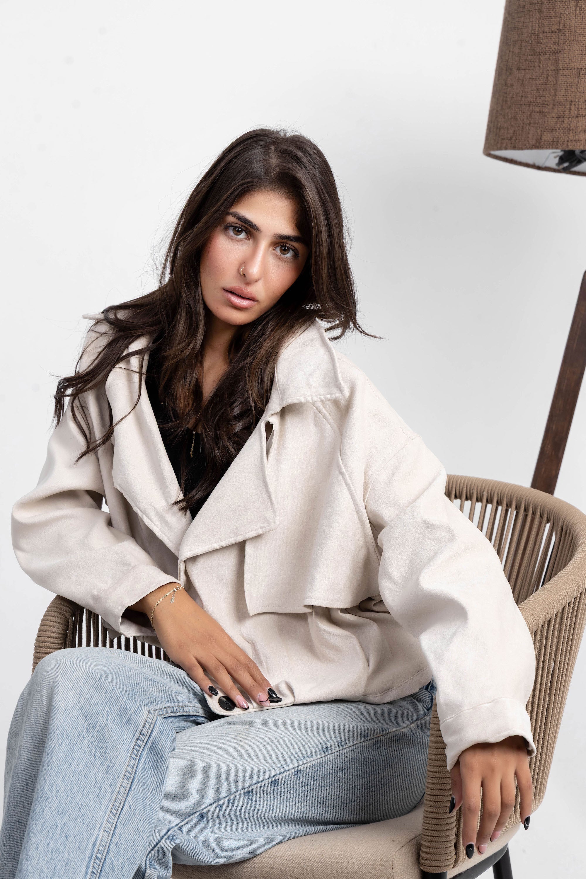 Woman sitting on a chair wearing a beige trench coat and blue jeans against a white background
