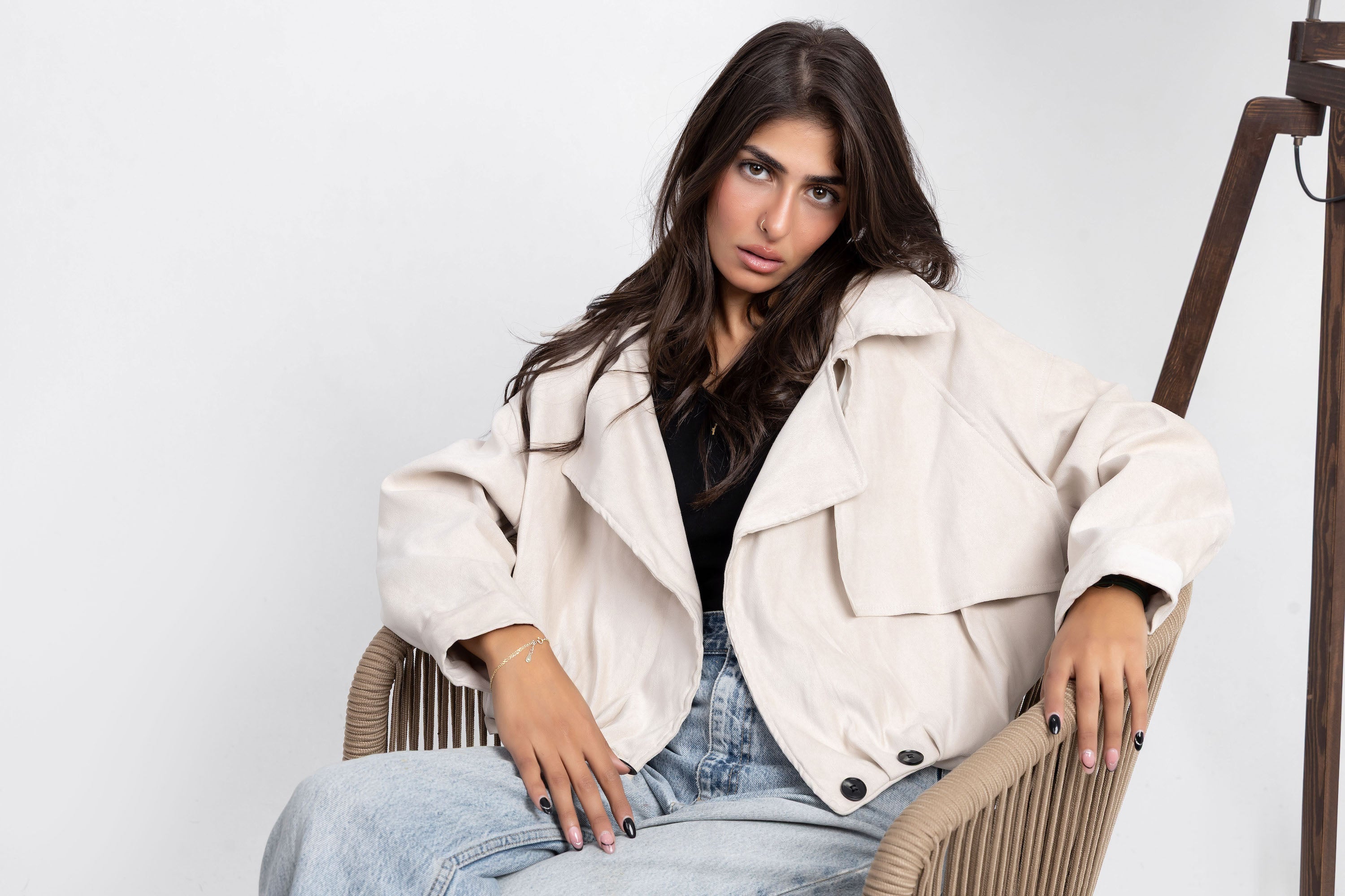 Woman sitting on a chair wearing a white jacket and blue jeans against a plain background