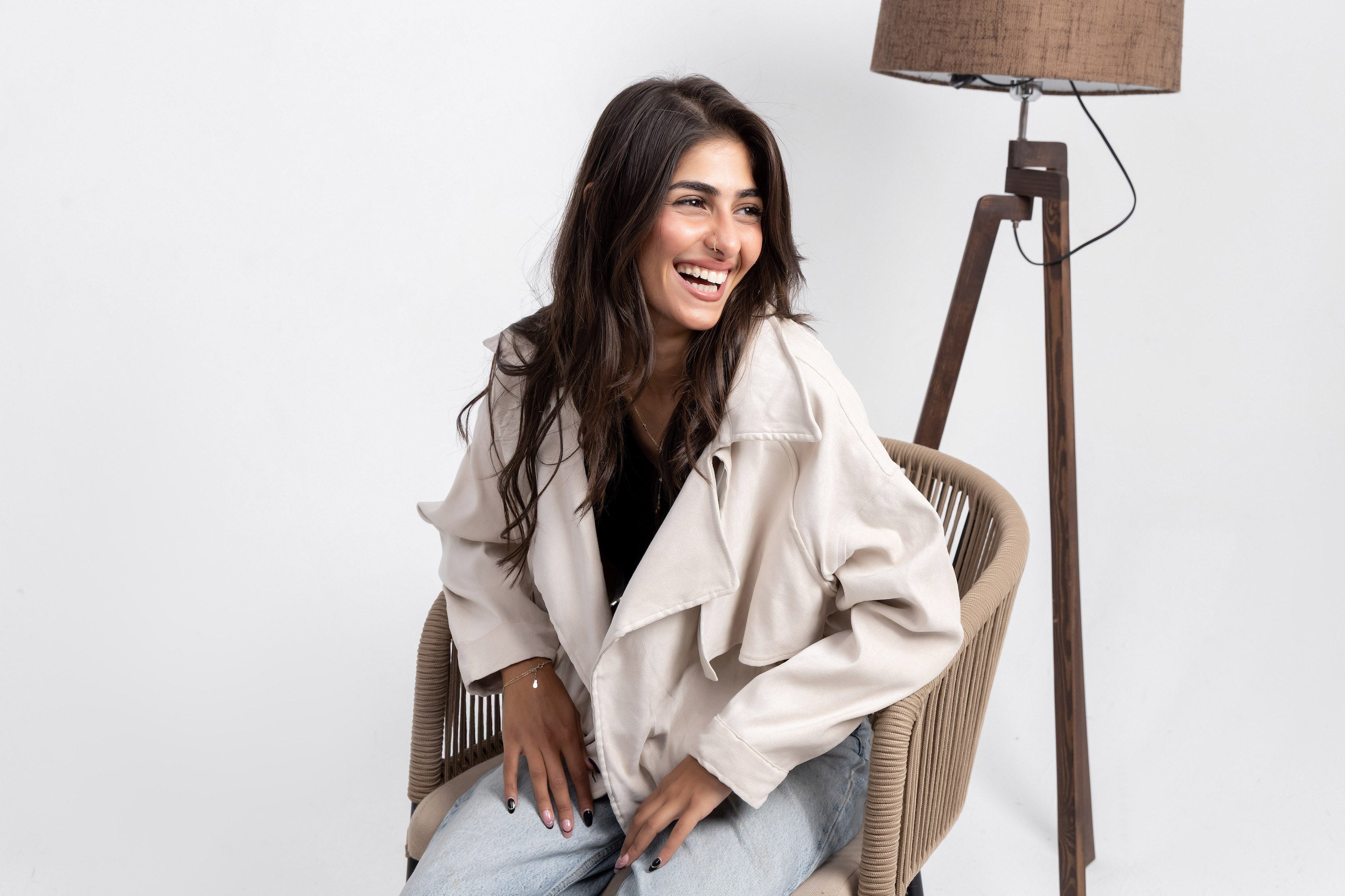 Woman sitting on a chair in a room with a lamp and white wall wearing a biege trench jacket